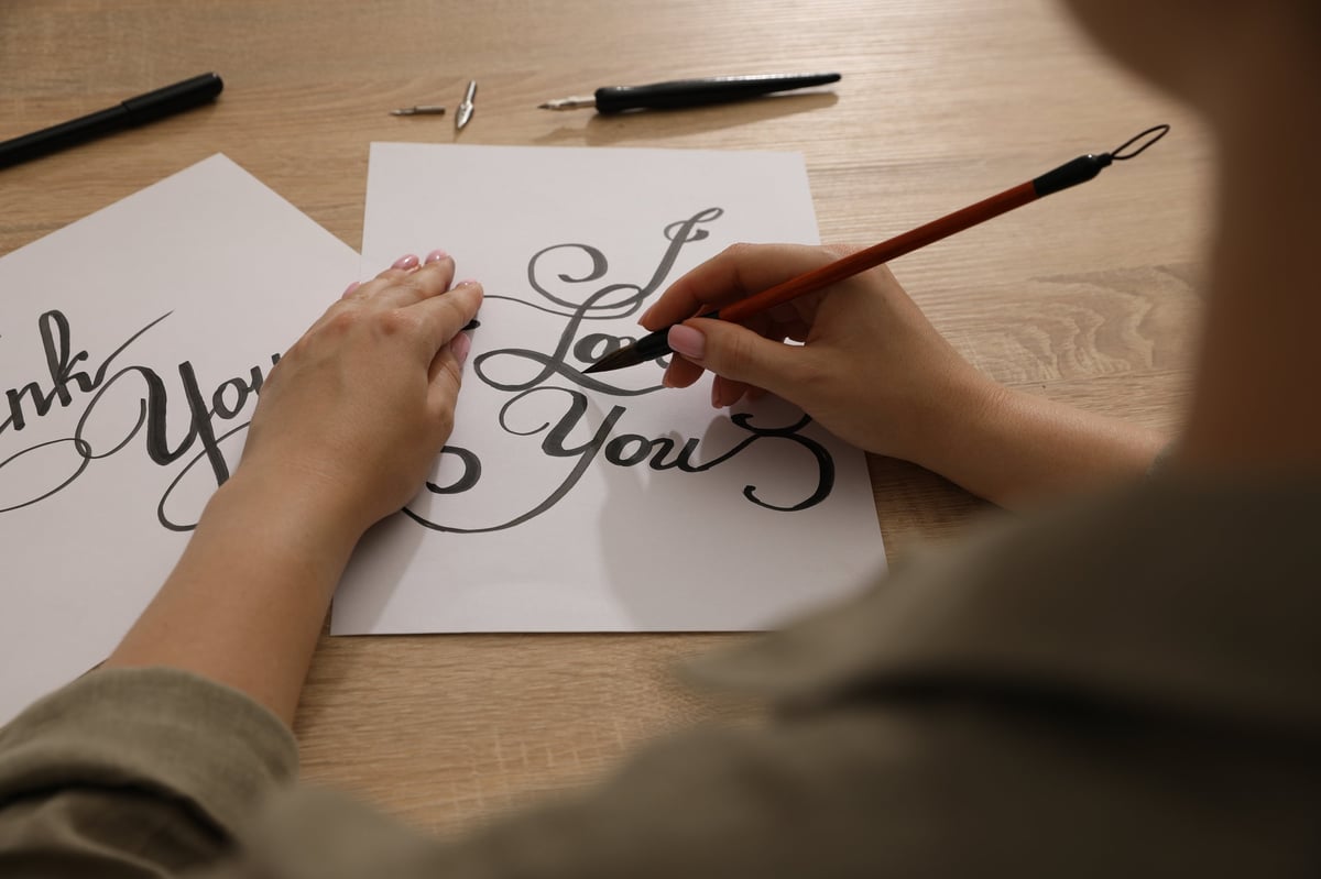 Calligraphy. Woman with brush writing phrase I Love You on sheet of paper at wooden table, closeup